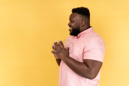 Photo for Side view portrait of cunning bearded man wearing pink shirt clasping hands and planning evil tricky prank or scheming, cheating with sly smile. Indoor studio shot isolated on yellow background. - Royalty Free Image