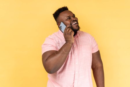 Foto de Portrait of cheerful man wearing pink shirt talking with friend on cell phone, looking away and smiling joyfully, having pleasant conversation. Indoor studio shot isolated on yellow background. - Imagen libre de derechos