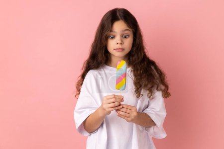 Téléchargez les photos : Portrait of shocked surprised little girl wearing white T-shirt holding delicious ice cream, looks at dessert with astonished expression. Indoor studio shot isolated on pink background. - en image libre de droit