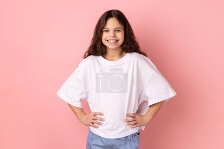 Photo for Portrait of delighted smiling little girl wearing white T-shirt standing hands on hips, looking at camera with happy expression. Indoor studio shot isolated on pink background. - Royalty Free Image