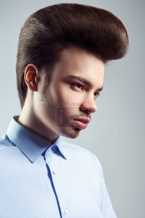 Téléchargez les photos : Side view portrait of handsome confident man with mustache and beard with retro classic elvis presley hairstyle, looking away, wearing blue shirt. Indoor studio shot isolated on gray background. - en image libre de droit