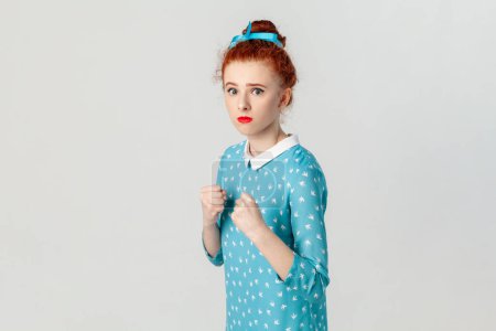 Foto de Portrait of confident serious red haired woman with hair bun, standing with clenched fists, being ready to attack, boxing, wearing blue dress. Indoor studio shot isolated on gray background. - Imagen libre de derechos