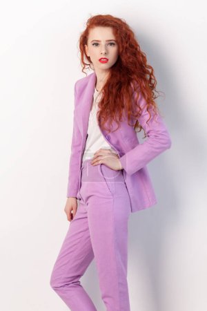 Foto de Portrait of attractive adorable confident ginger woman with curly hair, standing keeping hand on hip, looking at camera, wearing lilac suit. Indoor studio shot isolated on gray background. - Imagen libre de derechos