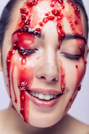 Téléchargez les photos : Closeup portrait of attractive woman fashion model with happy expression, smiling, keeps eyes closed, red jam flowing down on her face. Indoor studio shot isolated on gray background. - en image libre de droit