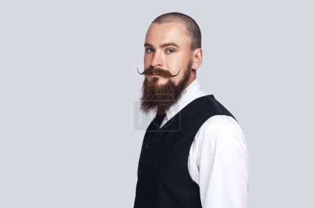 Téléchargez les photos : Side view portrait of serious attentive man with beard and mustache standing looking at camera with strict facial expression, being in bad mood. Indoor studio shot isolated on gray background. - en image libre de droit
