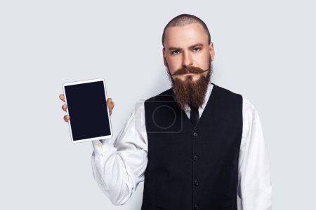 Téléchargez les photos : Portrait of serious assertive handsome man with beard and mustache showing tablet with empty display, advertisement area, copy space for promotion. Indoor studio shot isolated on gray background. - en image libre de droit