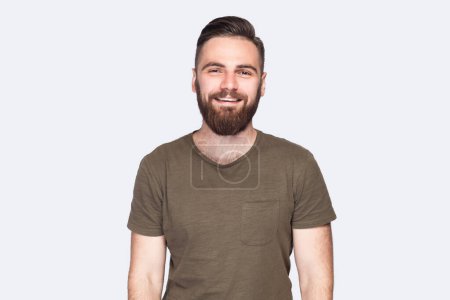 Téléchargez les photos : Portrait of handsome bearded man with stylish hairstyle wearing dark green T-shirt standing looking at camera with positive and optimism. Indoor studio shot isolated on light gray background. - en image libre de droit