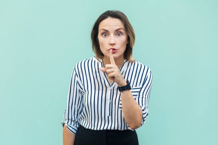 Photo for It's a secret. Portrait of serious middle aged woman wearing striped shirt standing with finger near mouth, showing shh gesture. Indoor studio shot isolated on light blue background. - Royalty Free Image