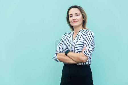 Foto de Portrait of confident middle aged woman wearing striped shirt standing with folded hands and looking at camera, being proud, keeps arms crossed. Indoor studio shot isolated on light blue background. - Imagen libre de derechos