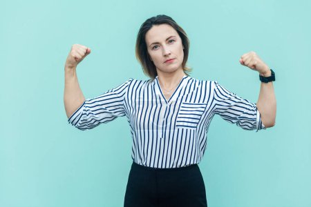 Photo for Portrait of strong confident attractive middle aged woman wearing striped shirt raised her arms, showing her power, looking at camera. Indoor studio shot isolated on light blue background. - Royalty Free Image