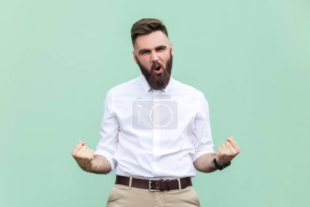 Téléchargez les photos : Portrait of excited bearded businessman wearing white shirt standing with clenched fists and screaming loud, celebrating victory and success. Indoor studio shot isolated on light green background. - en image libre de droit