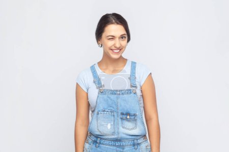 Photo for Portrait of positive playful young adult woman wearing denim overalls standing with toothy smile, winking and looking at camera, flirting. Indoor studio shot isolated on gray background. - Royalty Free Image