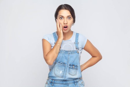 Photo for Surprised woman in denim overalls having stunned and shocked look, with mouth fell open and jaw dropped, listening to friend's story in full disbelief. Indoor studio shot isolated on gray background. - Royalty Free Image