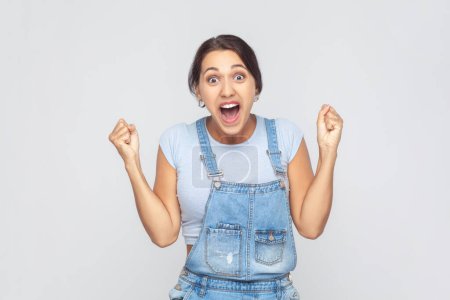 Photo for I am champion. Portrait of victorious delighted woman wearing denim overalls clenching fists, shouting for joy, screaming celebrating win success. Indoor studio shot isolated on gray background. - Royalty Free Image