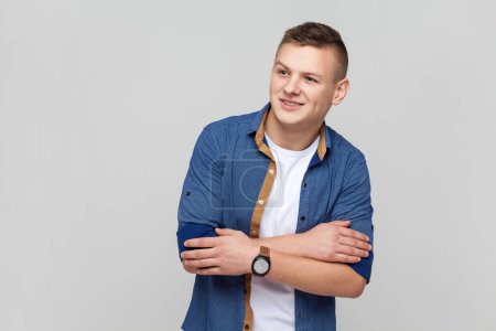 Photo for Portrait of handsome attractive young smiling teenager boy wearing blue shirt keeps hands folded, looking away with dreaming expression. Indoor studio shot isolated on gray background. - Royalty Free Image