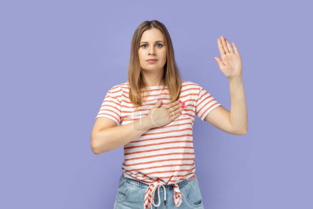 Téléchargez les photos : Portrait of serious patriotic blond woman wearing striped T-shirt raising one arm and putting on chest another making oath, swearing. Indoor studio shot isolated on purple background. - en image libre de droit