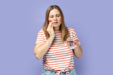 Téléchargez les photos : Portrait of sick blond woman wearing striped T-shirt holding cup in hands, having tooth pain after drinking cold or hot beverage. Indoor studio shot isolated on purple background. - en image libre de droit