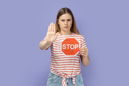 Téléchargez les photos : Portrait of blond woman wearing striped T-shirt holding red stop symbol and swing gesture with palm, forbidden sign with hand. Indoor studio shot isolated on purple background. - en image libre de droit