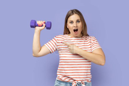 Téléchargez les photos : Portrait of amazed excited athletic blond woman wearing striped T-shirt holding dumbbell in hand and pointing at her biceps, likes to workout. Indoor studio shot isolated on purple background. - en image libre de droit