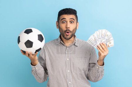 Téléchargez les photos : Shocked astonished businessman holding soccer ball and fan of dollar bills, sports betting, big win, looking at camera, wearing striped shirt. Indoor studio shot isolated on blue background. - en image libre de droit