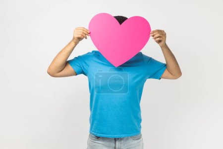Téléchargez les photos : Portrait of romantic unknown anonymous man wearing blue T- shirt standing holding big pink heart in front his face, expressing fondness and love. Indoor studio shot isolated on gray background. - en image libre de droit