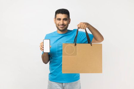 Téléchargez les photos : Portrait of pleased positive handsome man wearing blue T- shirt standing holding shopping bag and mobile phone with blank screen for advertisement. Indoor studio shot isolated on gray background. - en image libre de droit