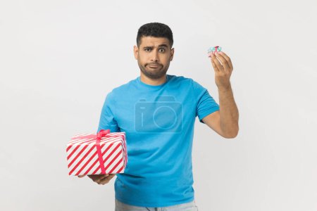 Téléchargez les photos : Portrait of puzzled attractive man wearing blue T- shirt standing choosing between two present boxes, choosing little gift box with disappointment. Indoor studio shot isolated on gray background. - en image libre de droit