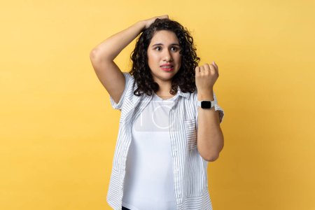 Téléchargez les photos : Portrait of beautiful adorable confused woman with dark wavy hair standing with puzzled facial expression, looks pensive, showing smart watch. Indoor studio shot isolated on yellow background. - en image libre de droit