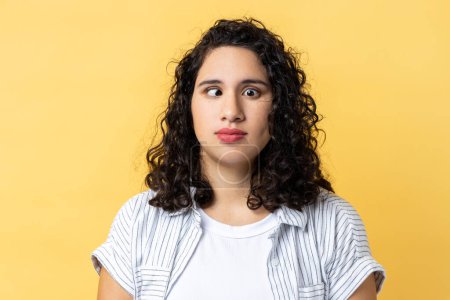 Foto de Portrait of funny woman with dark wavy hair making silly comical face with eyes crossed, thinking intensely looking dumb and confused. Indoor studio shot isolated on yellow background. - Imagen libre de derechos