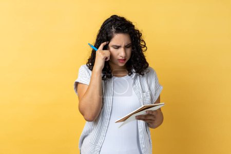 Foto de Portrait of thoughtful attractive young adult woman with dark wavy hair writing in paper notebook, journalist thinking about new article. Indoor studio shot isolated on yellow background. - Imagen libre de derechos