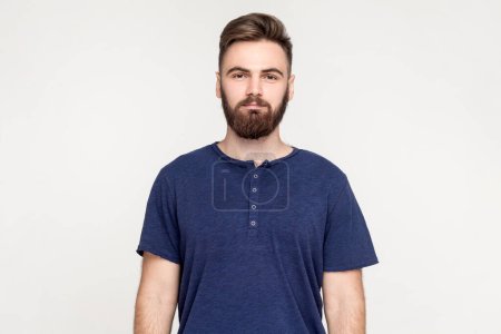 Foto de Portrait of serious handsome man with beard wearing dark blue T-shirt standing with hands down and looking at camera with strict expression. Indoor shot isolated on gray background. - Imagen libre de derechos