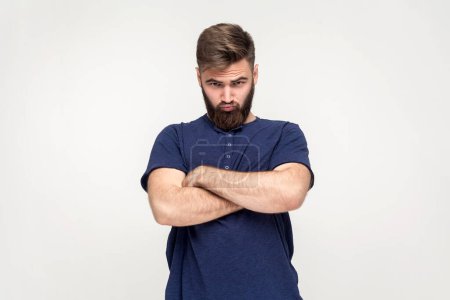 Photo for Portrait of man with beard wearing dark blue T-shirt being upset of bad news, looking at camera with frowning face, expressing sadness, keeps arms crossed. Indoor shot isolated on gray background. - Royalty Free Image