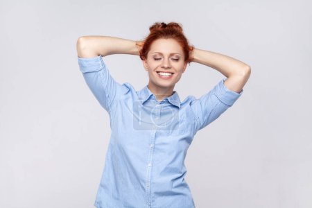 Photo for Portrait of beautiful attractive satisfied ginger woman wearing blue shirt standing with raised arms and closed eyes, smiling toothily. Indoor studio shot isolated on gray background. - Royalty Free Image