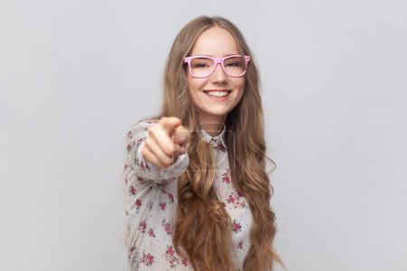 Photo for Portrait of pretty cheerful young adult woman in glasses with wavy blond hair looking at camera with toothy smile, pointing finger, suspecting you. Indoor studio shot isolated on gray background. - Royalty Free Image