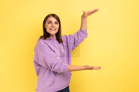 Téléchargez les photos : Portrait of happy charming woman presenting copy space between hands, holding empty place for commercial text, wearing purple hoodie. Indoor studio shot isolated on yellow background. - en image libre de droit