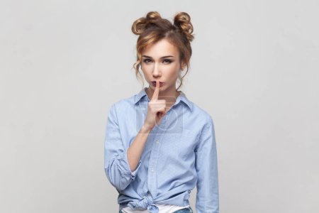 Photo for Portrait of strict blonde woman making hush gesture, pressed index finger to lips, looking at camera with serious expression, wearing blue shirt. Indoor studio shot isolated on gray background. - Royalty Free Image