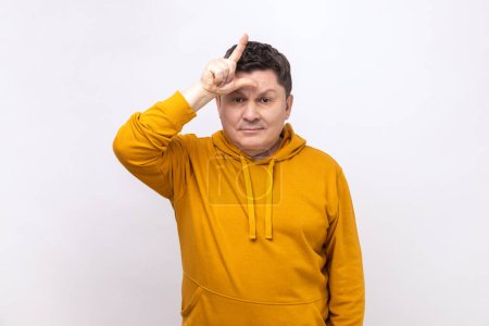 Photo for Gloomy depressed man showing looser gesture holding fingers near forehead, sad because of silly mistake, wearing urban style hoodie. Indoor studio shot isolated on white background. - Royalty Free Image