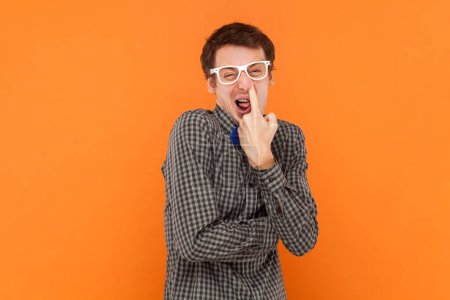 Téléchargez les photos : Portrait de l'homme nerd mettre le doigt dans son nez, plaisanter, mauvaises habitudes, comportement irrespectueux, porte une chemise avec un noeud papillon bleu et des lunettes blanches. Studio intérieur tourné isolé sur fond orange - en image libre de droit