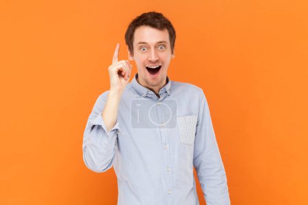 Photo for Portrait of inspired surprised man standing with raised finger, having sudden smart idea, looking at camera with open mouth, wearing light blue shirt. Indoor studio shot isolated on orange background. - Royalty Free Image
