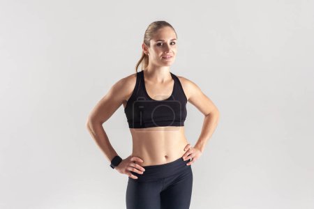 Photo for Self confident sporty blonde woman standing and looking at camera, keeps hands on hips, posing after workout, wearing black fitness clothing. Indoor studio shot isolated on gray background. - Royalty Free Image