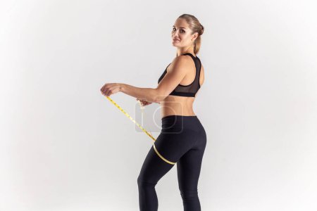 Téléchargez les photos : Side view portrait of delighted sporty blonde woman holding centimeter on her leg, checking result after sport marayhon, wearing black fitness clothing. Indoor studio shot isolated on gray background. - en image libre de droit