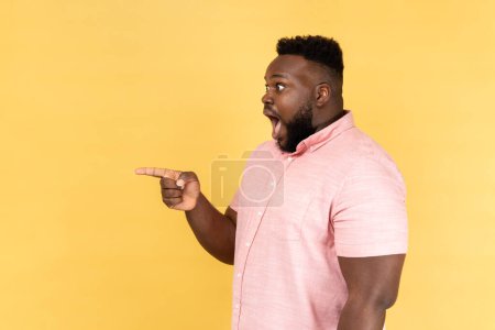 Photo for Side view portrait of shocked amazed man wearing pink shirt standing pointing away at copy space for advertisement or promotion. Indoor studio shot isolated on yellow background. - Royalty Free Image