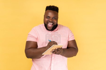 Photo for Portrait of man wearing pink shirt writing down in paper notebook, making to do list, having good mood, looking at camera with toothy smile. Indoor studio shot isolated on yellow background. - Royalty Free Image