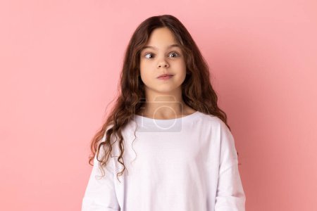 Téléchargez les photos : Portrait of little girl wearing white T-shirt crossing her eyes looking crazy and stupid, fooling around, having fun, vision problems. Indoor studio shot isolated on pink background. - en image libre de droit