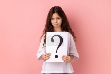 Photo for Portrait of winsome beautiful little girl wearing white T-shirt looking at camera, holding paper with question mark, thinks about tasks. Indoor studio shot isolated on pink background. - Royalty Free Image