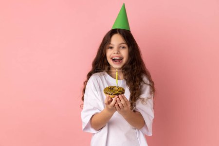 Téléchargez les photos : Excited little girl in green party cone on head holding cake with candle and looking at camera with excitement, expressing happiness. Indoor studio shot isolated on pink background. - en image libre de droit
