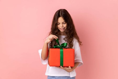 Foto de Retrato de una niña pequeña con una camiseta blanca abriendo una caja de regalo, desenvolviendo un regalo de cumpleaños para celebrar las vacaciones, tirando de la cinta, mirando el regalo. Estudio interior plano aislado sobre fondo rosa. - Imagen libre de derechos