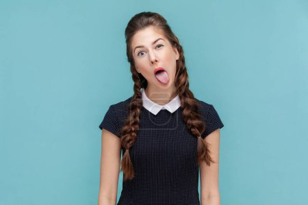 Téléchargez les photos : Portrait of naughty woman with braids sticking out tongue, making funny grimace, looking and winking at camera, wearing black dress. woman Indoor studio shot isolated on blue background. - en image libre de droit