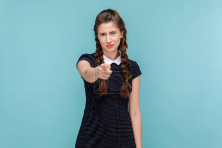 Foto de Portrait of funny beautiful young woman with braids indicating at you with index finger with frowning face, choosing you, wearing black dress. woman Indoor studio shot isolated on blue background. - Imagen libre de derechos
