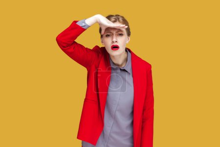Photo for Portrait of attentive concentrated woman with red lips standing looking far, keeps hand near her forehead, sees something far away, wearing red jacket. Indoor studio shot isolated on yellow background - Royalty Free Image